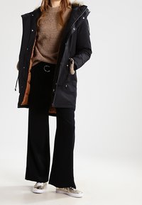 Parka noire avec une capuche doublée de fourrure, une doublure orange et de grandes poches frontales. Associée à un pull en maille marron et un pantalon noir à jambes larges.