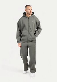 Grå hættejakke med lynlås og rummelig hætte, forlommer og matchende bukser. Materialet virker blødt med en glat tekstur. Hvide sneakers.