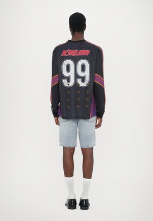 Black long-sleeve jersey with red and purple accents, large white number 99, and cross patterns; paired with light blue denim shorts.