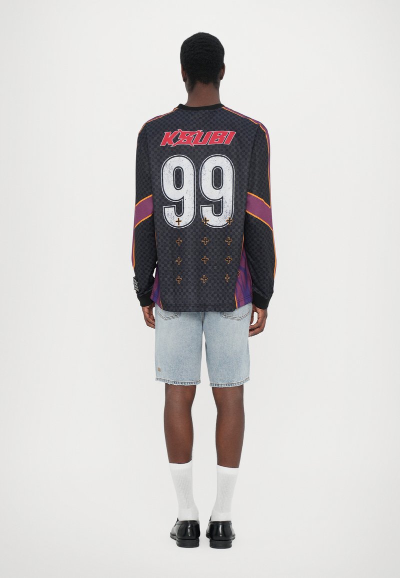 Schwarzes langärmeliges Trikot mit roten und lila Akzenten, große weiße Nummer 99 und Kreuzmuster; kombiniert mit hellblauen Denim-Shorts.