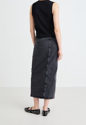 Woman wearing a black sleeveless top, high-waisted dark gray denim midi skirt, and black studded flat shoes, standing on a white floor.