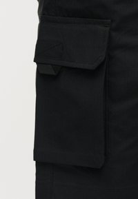 Μαύρη υφασμάτινη τσέπη cargo με καπάκι και ραμμένες λεπτομέρειες σε μαύρο ένδυμα, πιθανώς παντελόνι ή σορτς.