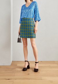 Top blu con paillettes e scollo a V, abbinato a una minigonna a quadri verde e blu. Scarpe nere con tacco a blocco, in mano una piccola borsa nera.