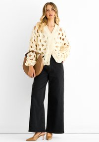 Cream knit cardigan with open hole pattern, five buttons, and dropped shoulders, paired with high-waisted black wide-leg jeans and beige heels.