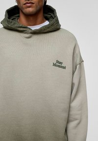 Olivengrüner und hellbeiger übergroßer Hoodie aus weichem Stoff. Mit einer Kängurutasche und dem Aufdruck "Stay Moment" in dunkelgrün.