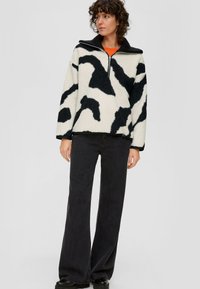 Kobieta ubrana w polarowy sweter w czarno-białe wzory z zamkiem błyskawicznym oraz czarne szerokie spodnie, stoi na tle jasnym.