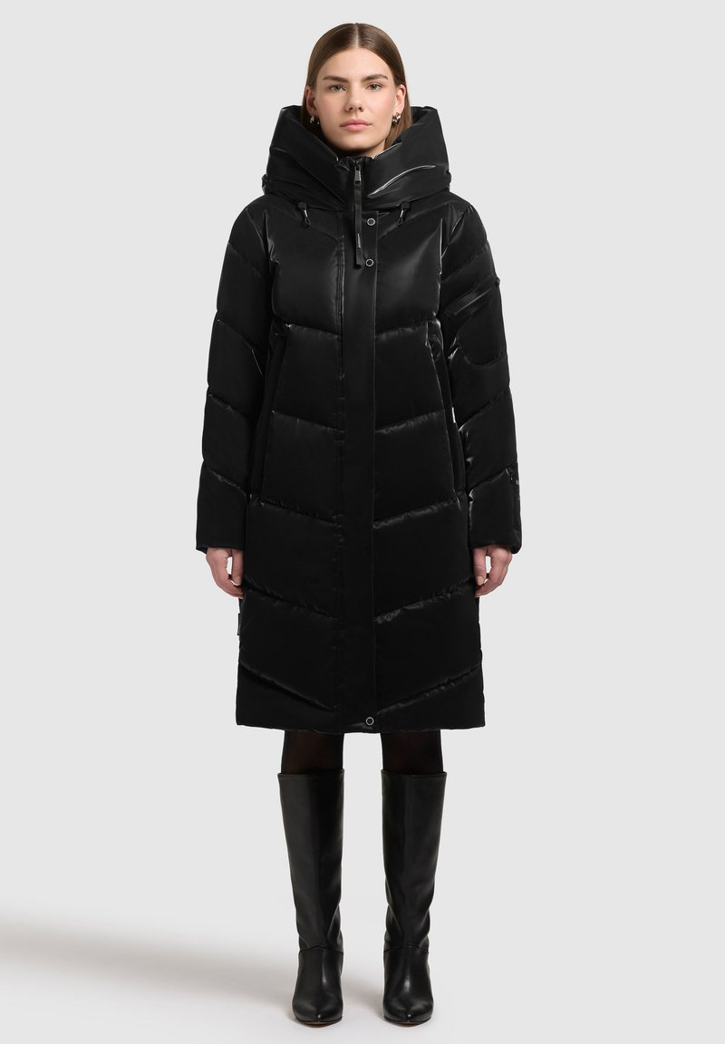 Sort polstret frakke med høj krave og stor hætte. Har en lynlås foran, sidelommer og chevron-quiltet mønster. Sammen med knæhøje støvler.