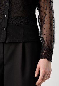 Blouse noire avec des détails en dentelle, manches transparentes à pois, boutonnière sur le devant et poignets en dentelle élaborés, assortie à un pantalon noir.