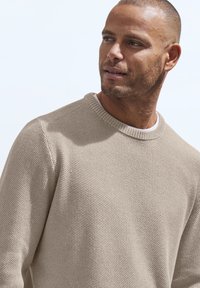 Beige gestrickter Pullover mit Rundhalsausschnitt und strukturiertem Muster. Der Stoff wirkt weich und bequem, ideal für lässige Outfits.