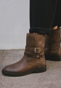 Bottes courtes en cuir marron avec un bout arrondi, des accents de boucle et une doublure en fausse fourrure en haut. Semelle plate, fermeture éclair à l'arrière.