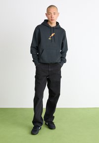 Kulforklæde med frontlomme og gul grafik, parret med sorte straight-leg jeans og sorte sneakers. Ensfarvet baggrund.