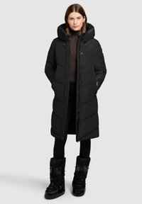 Frau in einem langen schwarzen gepolsterten Wintermantel mit Kapuze, schwarzen Leggings und schwarzen geschnürten Schneestiefeln, die vor einem schlichten Hintergrund steht.