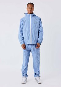 Cazadora ligera de color azul claro con costuras blancas en contraste, cremallera completa y capucha, combinada con pantalones a juego en una tela suave y ligera.