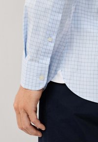 Mannelijke hand rustend aan de zijkant, gekleed in een lichtblauw geruit overhemd met lange mouwen, dat aan de manchetten is dichtgeknoopt, en donkere broek.