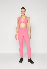 Rosa sports-bh med en vridd frontdesign, kombinert med høyt midjeførte rosa tights. Modellen har på seg svarte treningssko, bakgrunn grå.