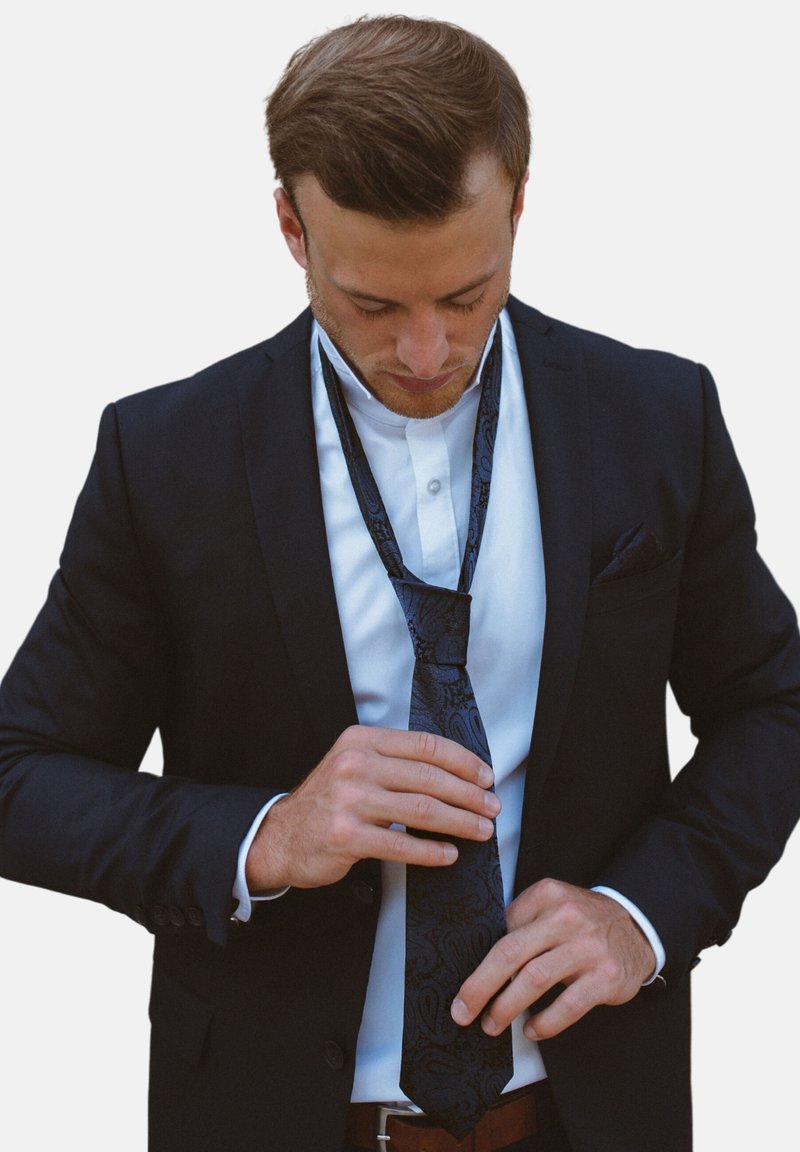 Cravate noire en paisley avec un motif texturé, portée sur une chemise blanche et sous une veste de costume noire, pendant que des mains ajustent la cravate. Ceinture marron visible.
