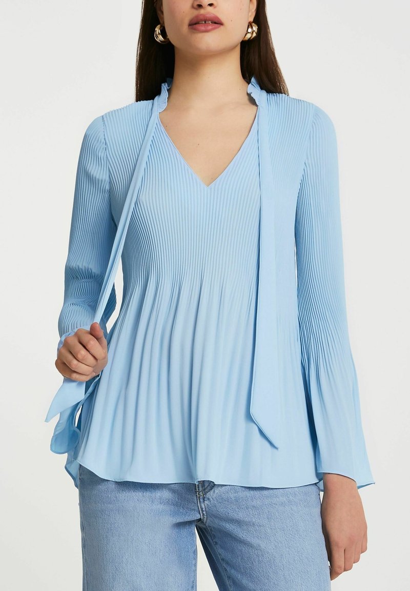 Femme portant un chemisier plissé bleu clair à manches longues, avec un col en V et un détail noué au col, associé à un jean bleu clair.