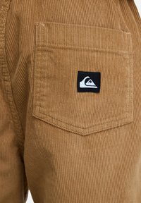 Poche arrière d'un pantalon en velours côtelé marron avec un écusson noir et blanc du logo Quiksilver cousu au centre supérieur de la poche.