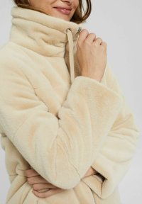 Femme portant une veste peluche beige clair, tenant le col près de son menton avec une main, souriant doucement, sur un fond uni.