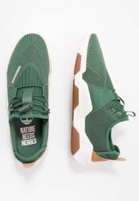 Baskets de sport vertes en tissu texturé avec un design à lacets, semelle intermédiaire blanche et accents marron au niveau du talon.