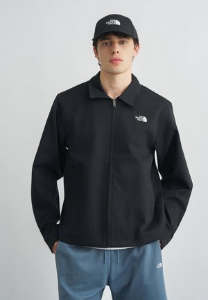 Jeune homme portant une veste zippée noire North Face et une casquette noire assortie, debout devant un fond clair uni.