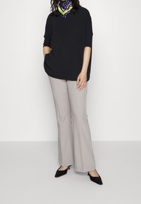 Schwarzes Oversized-Shirt mit einer Tasche, hellgraue weit geschnittene Hose und schwarze Low-Heel-Schuhe. Schal mit Knotenmuster um den Hals.