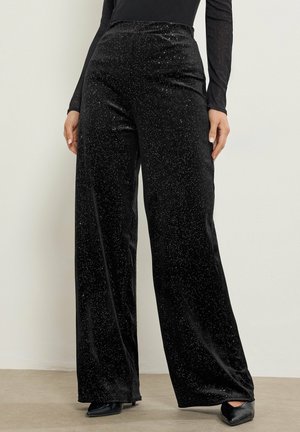 Frau trägt schwarze, weit geschnittene Hose mit hoher Taille und funkelnder Textur sowie schwarze spitze Schuhe, steht vor einer schlichten Wand.
