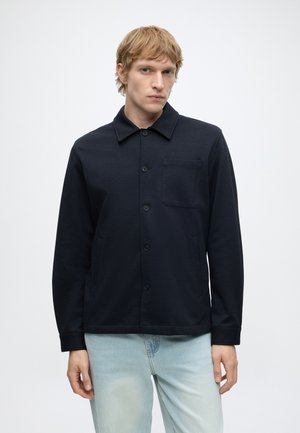 Lyshåret mand iført en knappet sort jakke med en brystlomme og lyseblå jeans, stående mod en ensfarvet hvid baggrund.