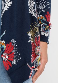 Mörkblå tröja med livfulla mönster av röda, gula, vita och flerfärgade blommor och blad, bärs med ljusblå jeans, handen vilar vid sidan.