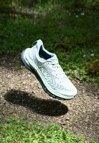 Vaaleanharmaa juoksukenkä, jossa on teksturoitu päällinen, verkko-osat, vihreät yksityiskohdat, pehmustettu pohja ja näkyvä brändilogo. Kelluu sorapinnan yläpuolella.