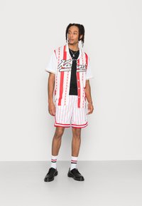 Rot-weißer gestreifter Kurzarm-Hemd, schwarzes T-Shirt darunter, passende gestreifte Shorts, schwarze Schuhe und weiße Socken mit roten Akzenten.