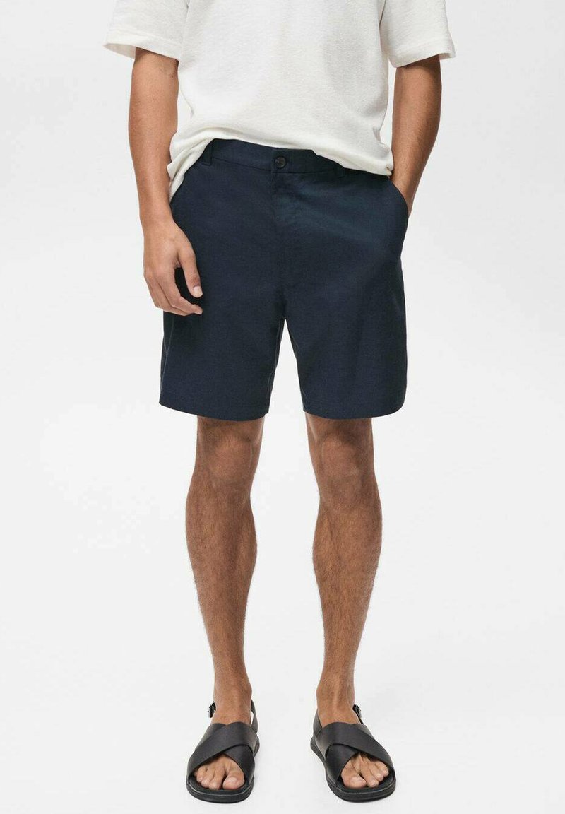 Pantaloni scurți din bumbac bleumarin cu croială dreaptă, buzunare laterale și închidere cu nasture în față, asortați cu sandale negre.
