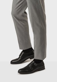 Zapatos de charol negros con doble hebilla, combinados con pantalones de vestir grises y calcetines negros, sobre un fondo claro.
