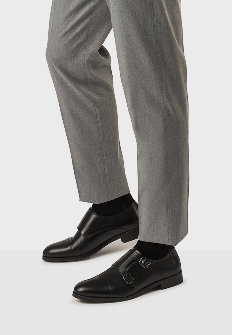 Zapatos de charol negros con doble hebilla, combinados con pantalones de vestir grises y calcetines negros, sobre un fondo claro.