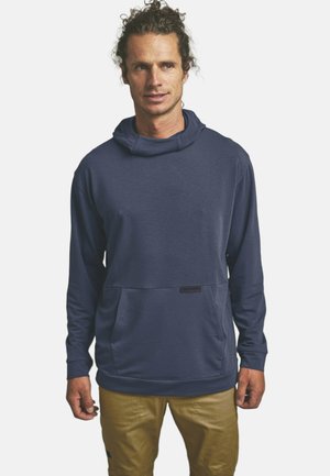 Mann mit lockigem Haar, der einen marineblauen Kapuzenpullover und beige Hosen trägt, vor einem schlichten weißen Hintergrund steht.