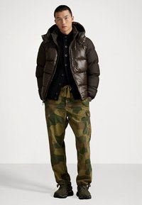 Brązowa puchowa kurtka z kapturem, czarna koszula zapinana na guziki, spodnie cargo w kamuflażu oraz ciemne trampki. Gładka faktura i luźny krój.