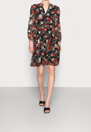 Kobieta ubrana w czarną sukienkę w kwiaty do kolan z długimi prześwitującymi rękawami i czarne czółenka z odkrytymi palcami, stojąca na jednolitej szarej podłodze.