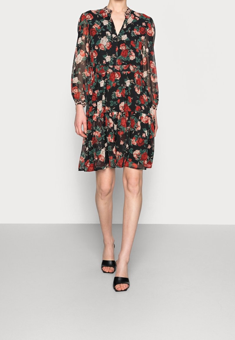 Femme portant une robe noire à fleurs longueur genou avec des manches longues transparentes et des talons ouverts noirs, debout sur un sol gris uni.