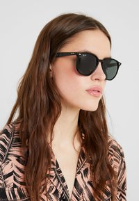 Sorte Ray-Ban solbriller med mørke linser, rund form og metal detaljer. Modellen har en lyserød skjorte med et sort bladmønster på.