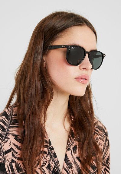 Червени слънчеви очила Ray-Ban с тъмни стъкла, закръглена форма и метални акценти. Моделът носи розова риза с черен листен речен.