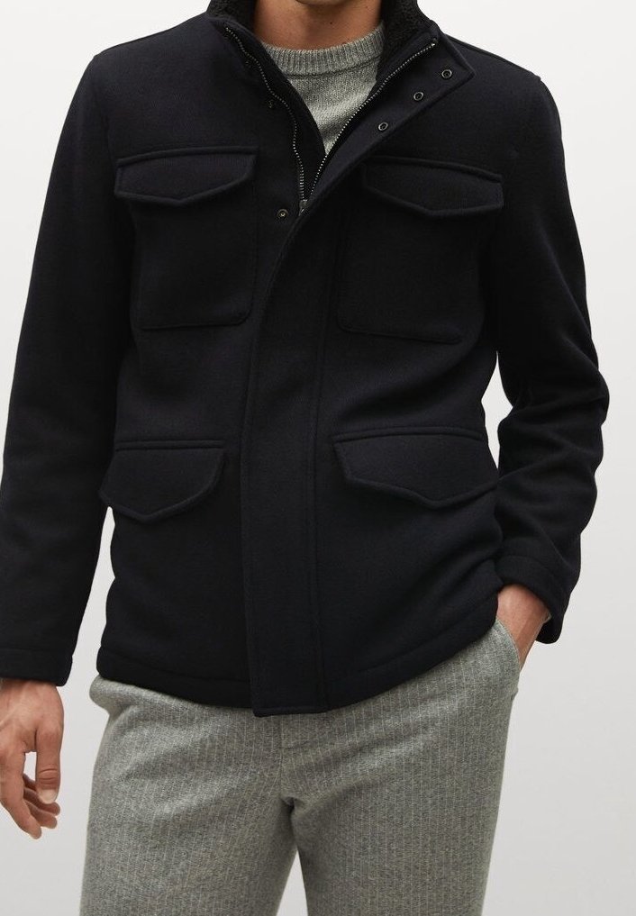 Homme portant une veste en laine noire boutonnée avec quatre poches à rabat sur un pull en maille gris et un pantalon gris texturé.