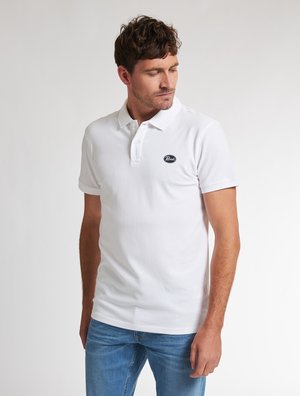 Weißes Polohemd aus Baumwolle mit einem Kragen mit drei Knöpfen und einem kleinen runden Logo auf der linken Brust. Getragen mit blauen Jeans.