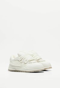 Baskets blanches avec un dessus synthétique lisse, une languette rembourrée, des lacets épais et une semelle épaisse dotée de rainures horizontales.