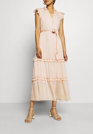Femme portant une robe midi sans manches rose clair avec des détails à volants, une ceinture nouée à la taille et des sandales à talons beiges avec des brides à la cheville.