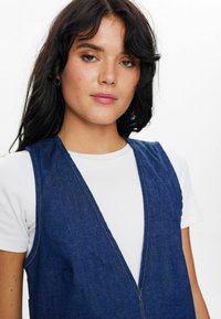 Denim jopič v temno modri barvi z globokim V-izrezom in strukturirano obliko, z kontrastnim svetlim šivom in stranskimi žepi, oblečen preko bele srajce.