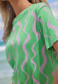 Mujer vestida con una camiseta verde con rayas onduladas de color púrpura claro, de pie al aire libre cerca de una playa con olas del océano de fondo.