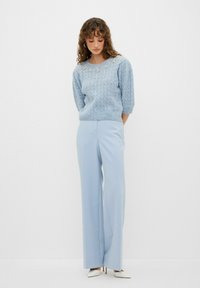 Maglione lavorato a maglia di colore blu chiaro con un motivo testurizzato e un'applicazione floreale sul collo, abbinato a pantaloni ampi blu chiaro e tacchi bianchi.