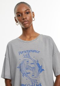 Camiseta gris de gran tamaño con un gráfico azul de dos peces, junto con el texto "Perseverancia y Buena suerte", en un tejido casual.
