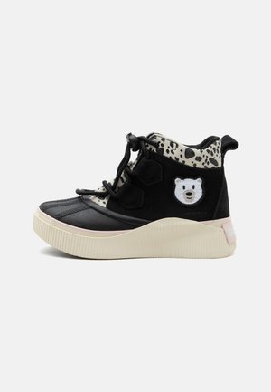 Zapato impermeable negro y crema para niño con cuello de estampado animal, cordón y correa ajustables, y parche con cara de oso blanco en el lateral.