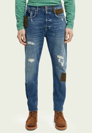 Mann trägt ausgewaschene blaue Jeans mit braunen Stoffflicken und hellbraunen Schnürstiefeln, kombiniert mit einem grünen Langarmshirt.